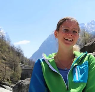 Smiling woman wearing green and blue hiking jacket with sunglasses on head, standing outdoors with mountain peaks and forested valley in background