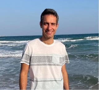 Man wearing striped white and gray shirt smiling on beach with ocean waves behind him
