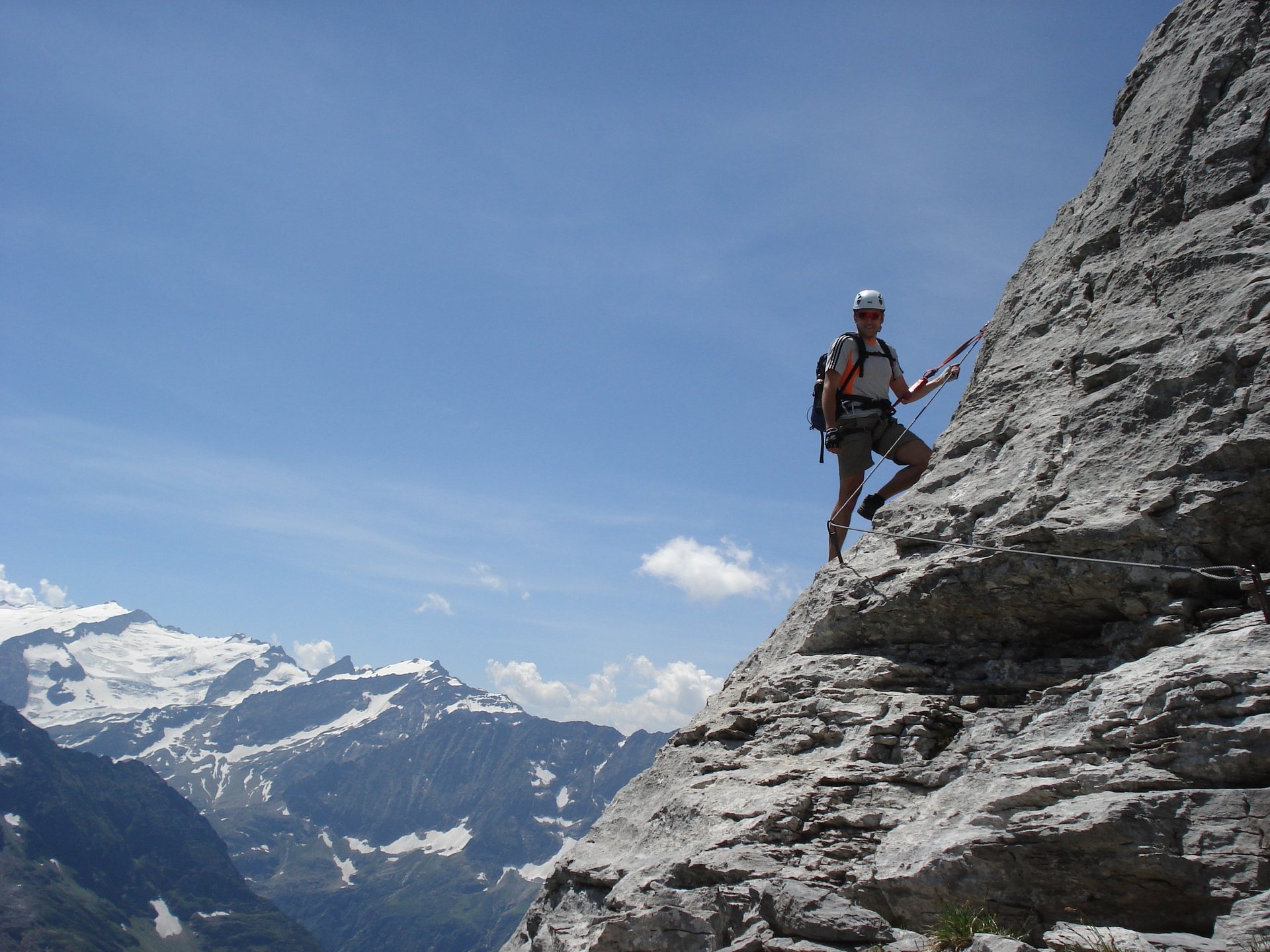 Rock climber rappelling down a steep gray cliff face with snow-capped alpine mountains in the background