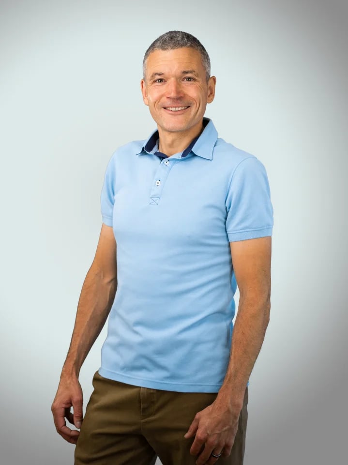 Man with short gray hair smiling at camera wearing light blue polo shirt and olive pants against plain white background