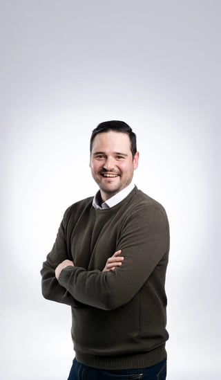 Professional headshot of a smiling man with dark hair wearing an olive green sweater over a collared shirt, arms crossed against a light background